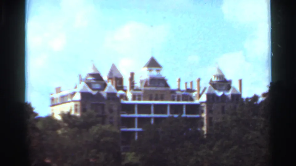 Visitor-shot 1965 8mm view of a large turreted building partly screened by trees under bright sky.