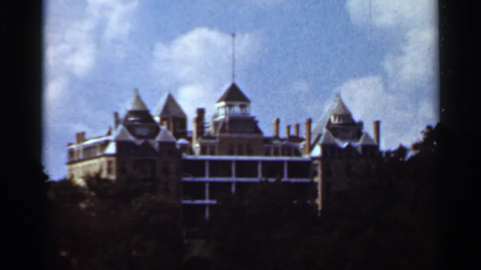 1965 home-movie frame of a castle-like building with turrets, balconies, and a flagpole above a wooded foreground.