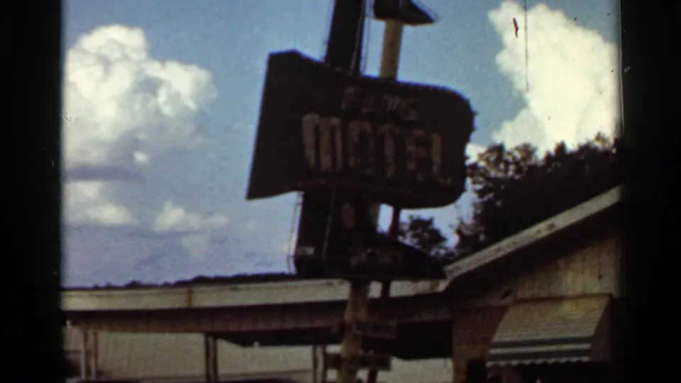 1960s 8mm frame of the Elms Motel sign, with the vacancy area and low motel roofline visible below the clouded sky.