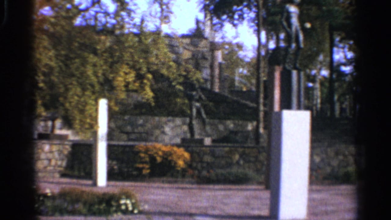Park entrance area with white pillars, stone wall, trees, shrubs, and a tall sculpture partly visible.