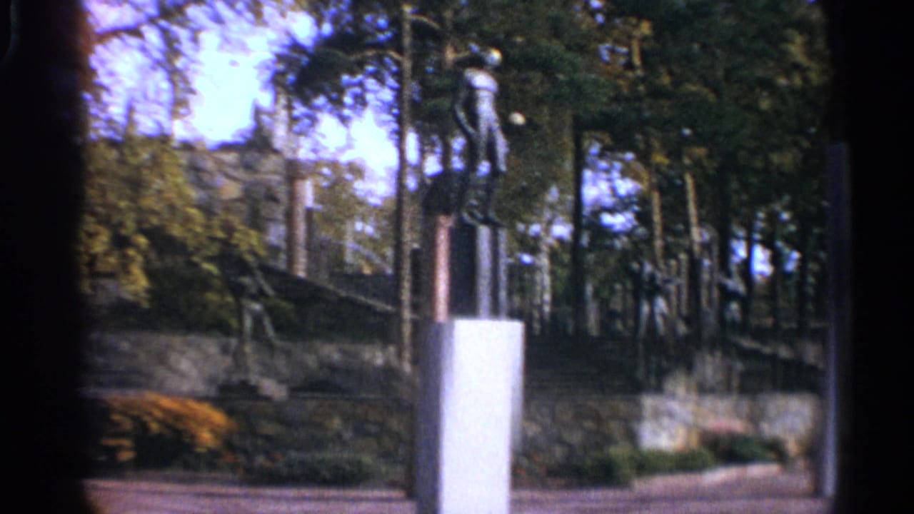 White pillar and stone wall beside a tree-lined park, with sculpture forms visible above the landscaping.