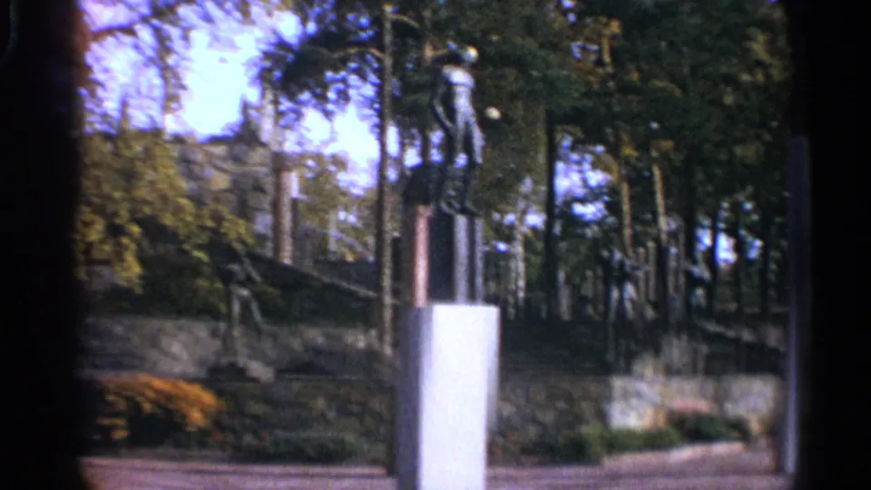 Leningrad park view from 1965 showing a white plinth, stone wall, trees, shrubs, and slender sculpture forms in the background.