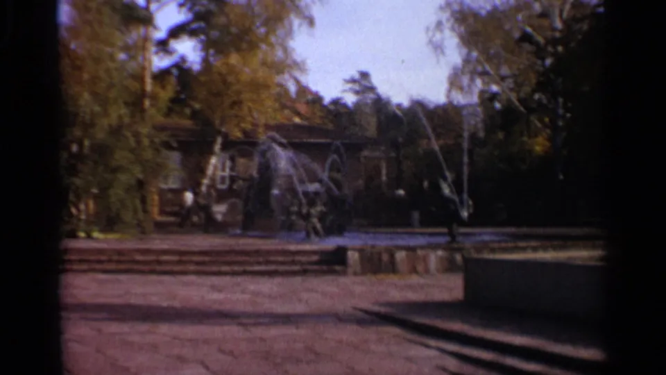 1965 Leningrad park scene with a fountain spraying across a paved area, trees, and a background structure.