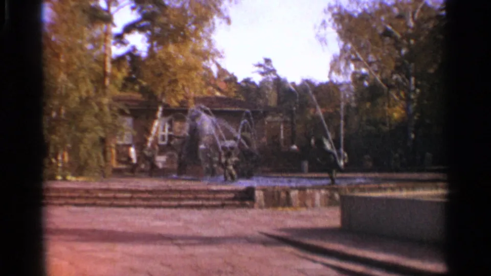 1965 Leningrad park scene with a fountain, paved open area, trees, and low structures in soft 8mm color.