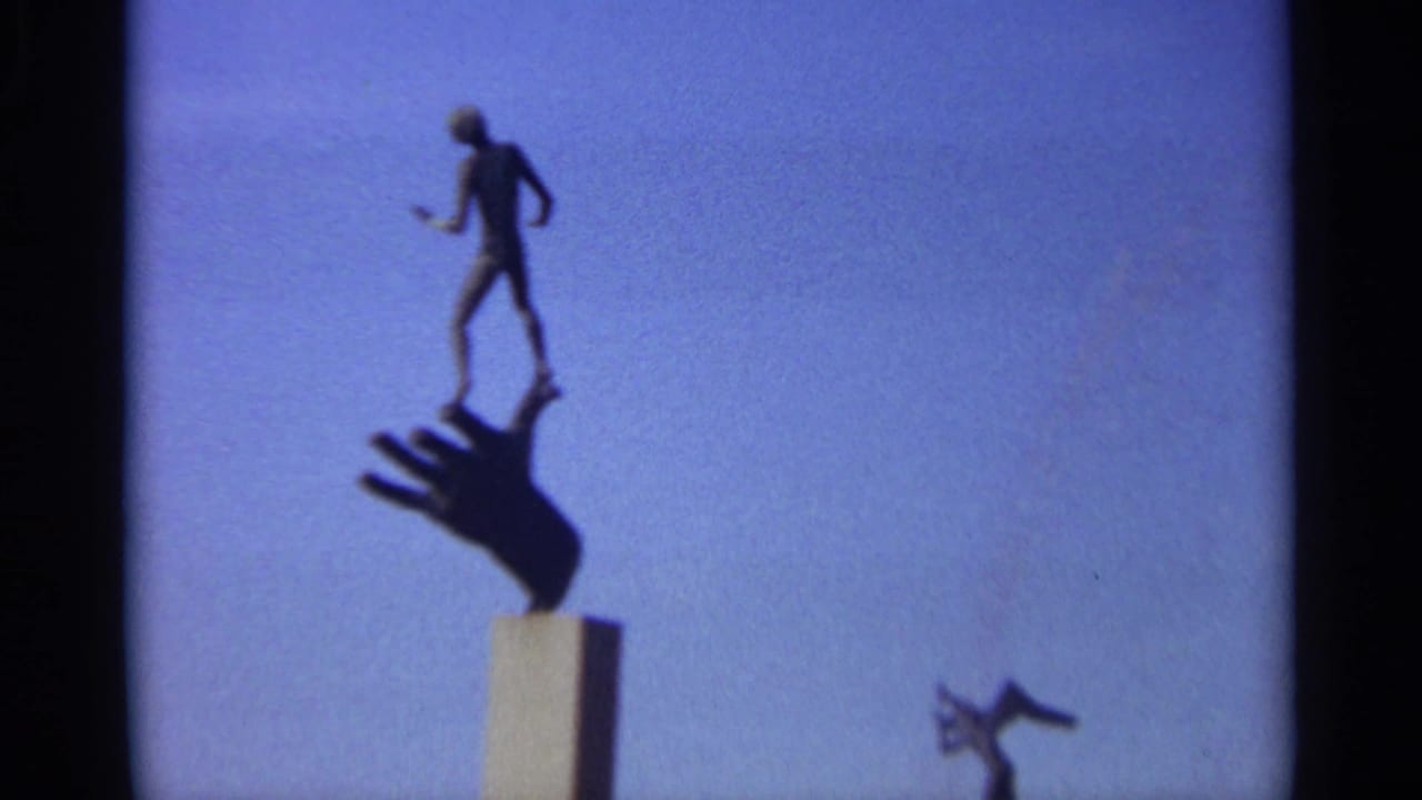Silhouetted sculpture of a figure standing on a large hand against blue sky.