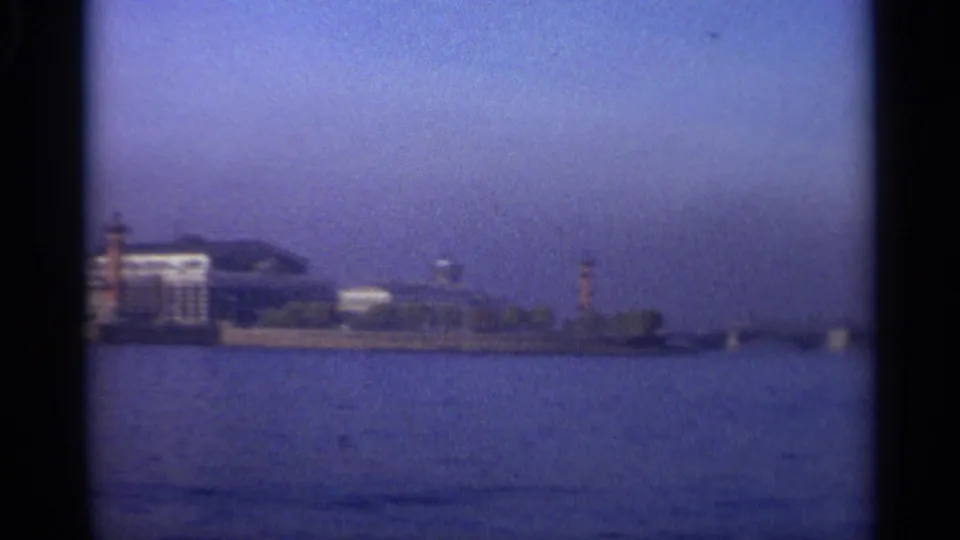 1965 Leningrad waterfront seen from across the water, with pale buildings, a tower, haze, and visible 8mm frame edges.