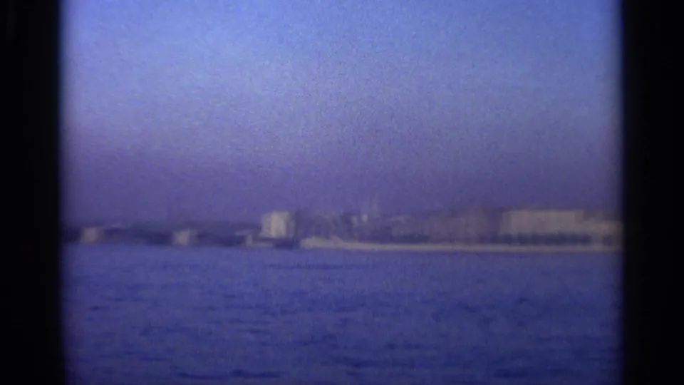 Distant Leningrad shoreline with a low bridge or pier-like line, pale buildings, and rippled water in a blue 8mm frame.