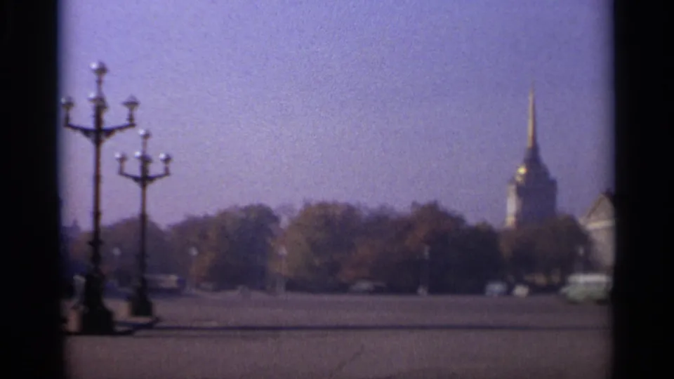 1965 Leningrad home-movie view across a broad square with ornate lamps, trees, a distant gilded spire, and sparse vehicles.