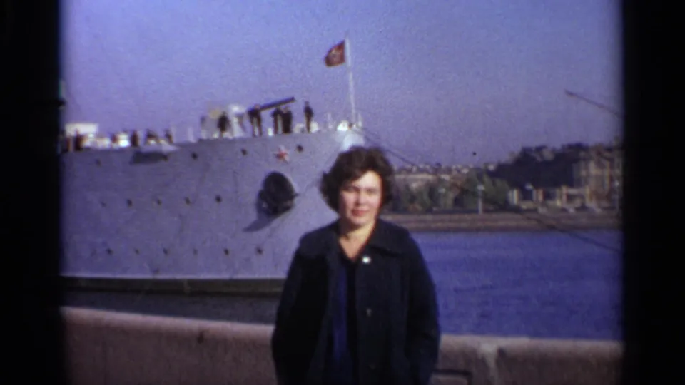 Woman poses on a Leningrad waterfront in 1965, with a gray Soviet warship, red flag, crew, and water behind her.