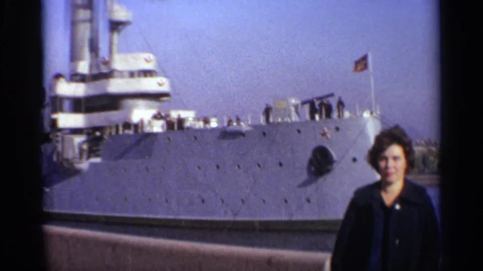 1965 Leningrad harbor frame with a woman in front of a gray Soviet warship displaying a red flag.