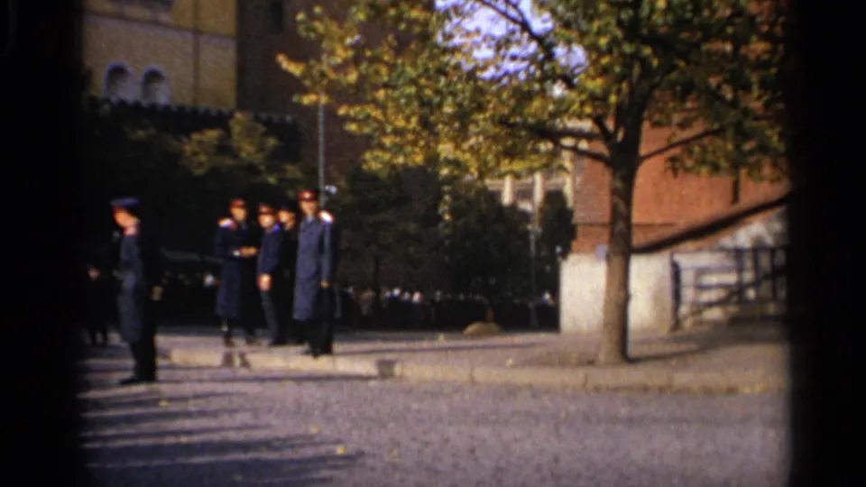 1965 Leningrad frame showing uniformed men standing outside near a tree-lined paved area and masonry building.