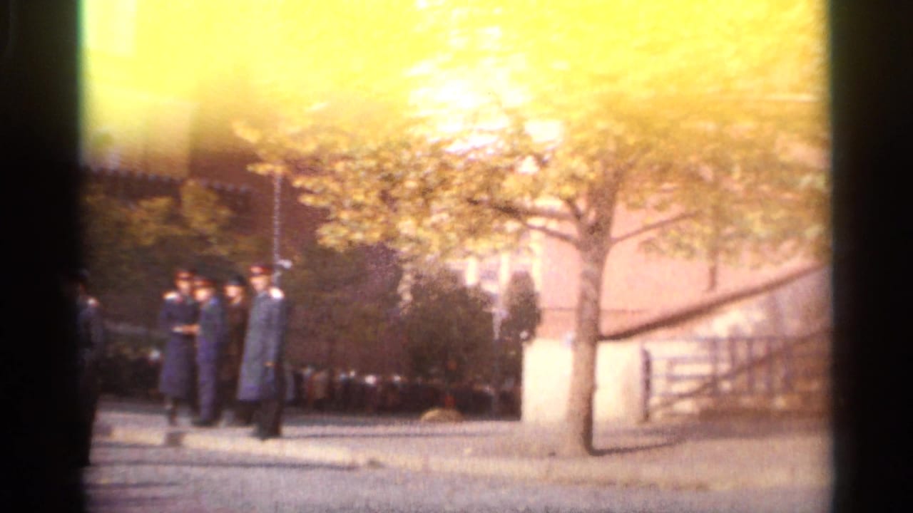 1965 Leningrad home-movie frame showing men, some in dark clothing, standing near a sidewalk and trees beside a large building.