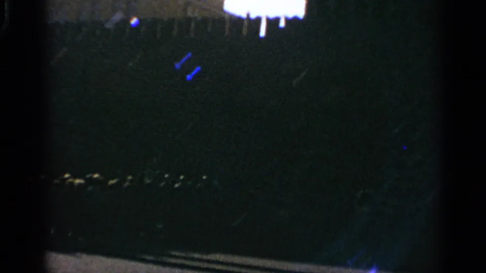 Dark underexposed 1965 home-movie frame with indistinct silhouettes, a bright upper edge, and small light marks near the bottom.