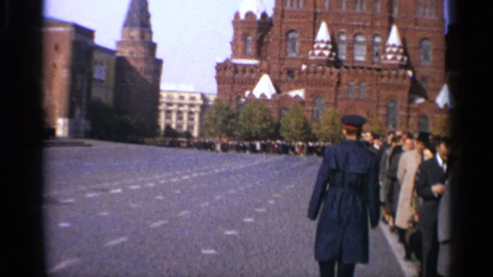 1965 Leningrad home-movie view of a broad square with historic red-brick buildings, a distant tower, crowds, and a uniformed figure in the foreground.