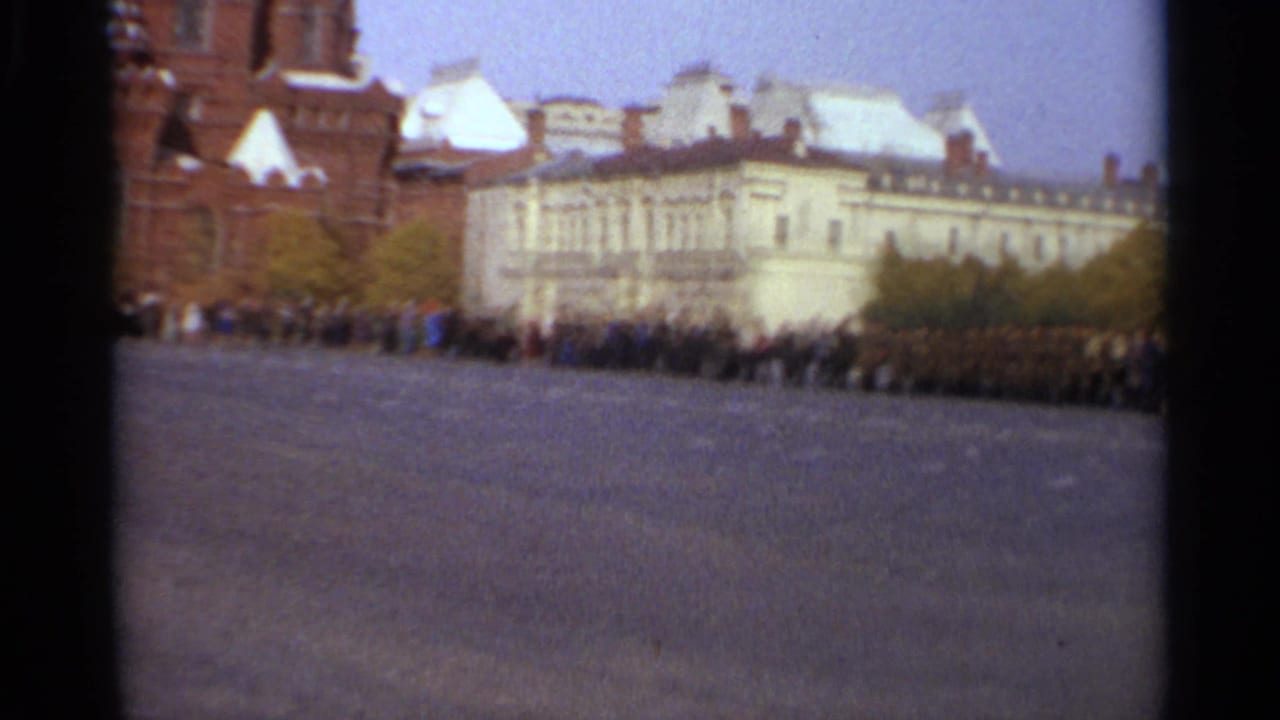 1965 home-movie frame of a distant crowd lined up near historic red-brick and pale buildings in a broad Soviet public square.
