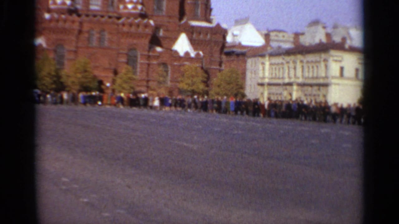 Distant crowd in a large public square, with red-brick historic architecture and a pale formal building visible behind them.