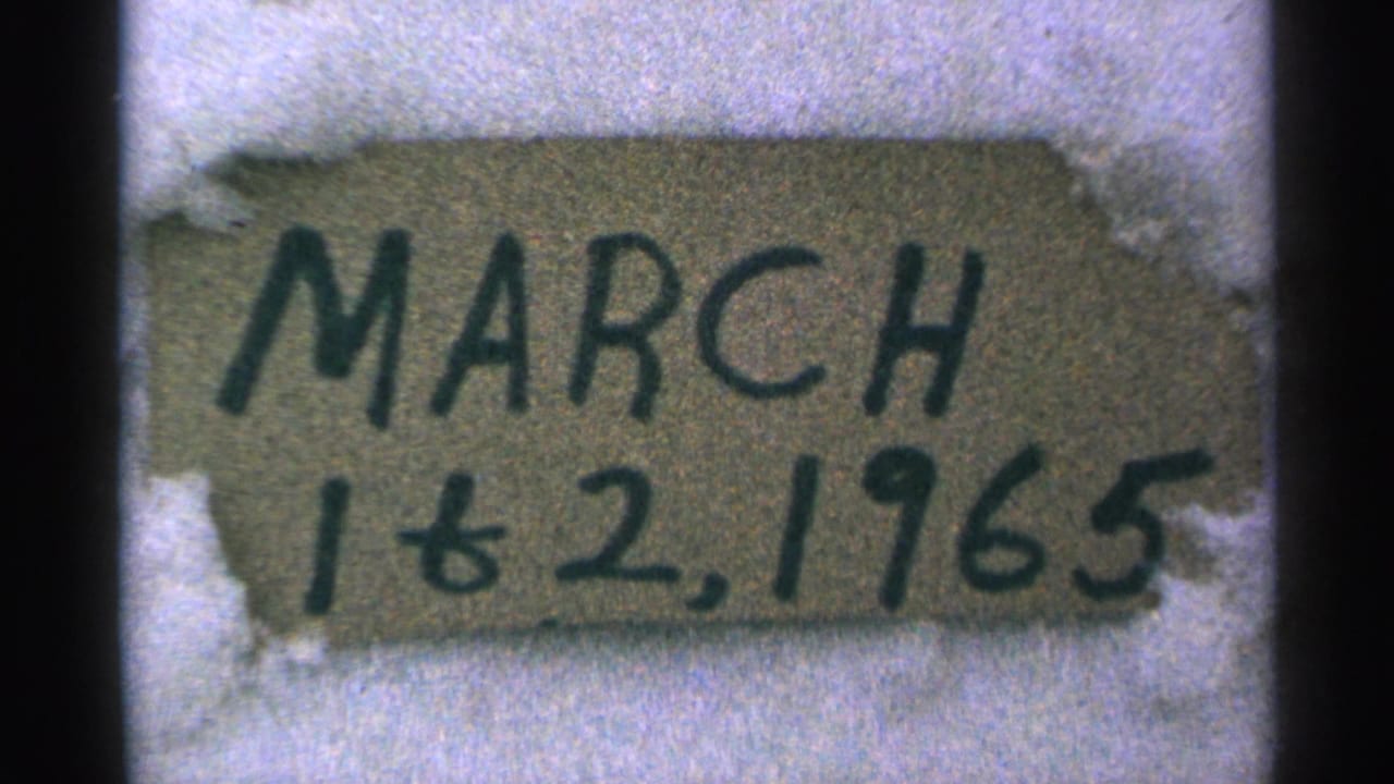 1965 Minnesota 8mm frame showing a close-up handmade cardboard date sign partly surrounded by snow.