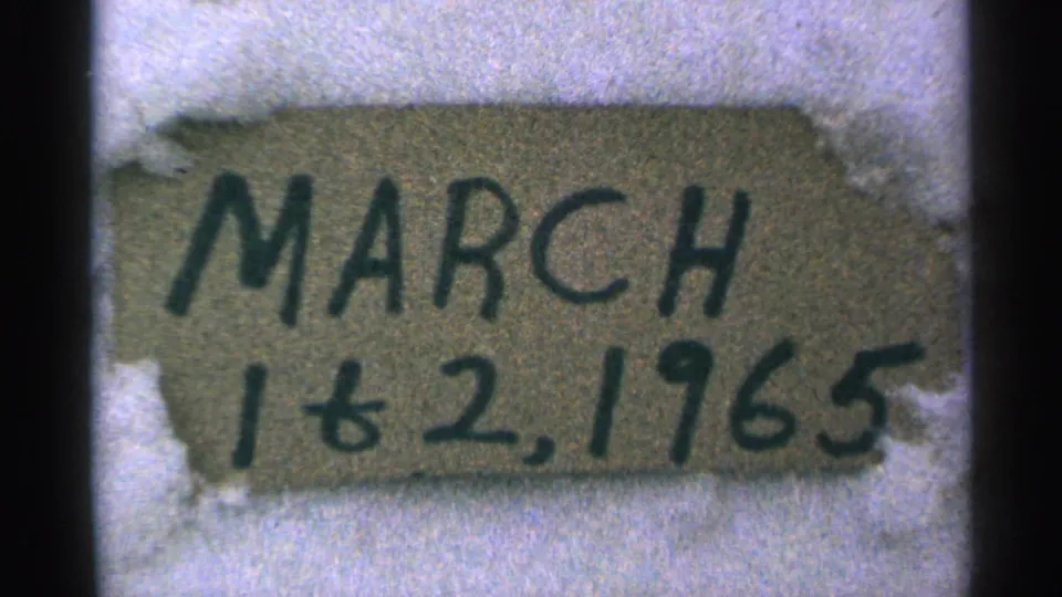 1965 Minnesota 8mm frame showing a close-up handmade cardboard date sign partly surrounded by snow.