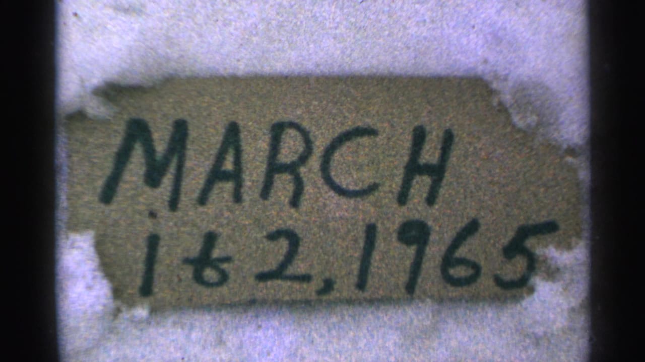 Close 8mm view of a handwritten March 1 & 2, 1965 sign in snow, with grain and soft focus visible.