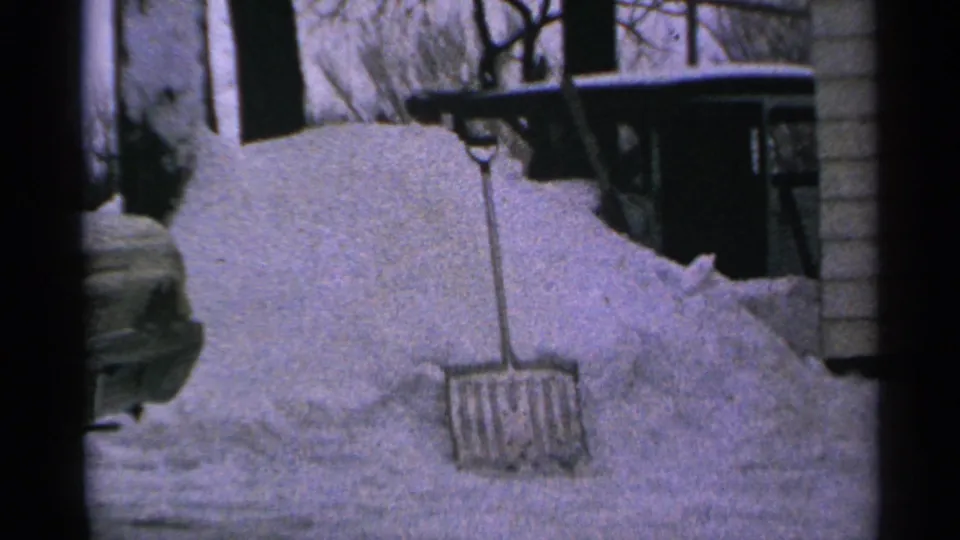 1965 Minnesota 8mm frame showing a snow shovel planted in a high snowbank near a house wall and porch area.
