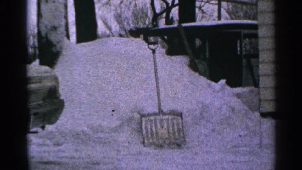 1965 Minnesota home-movie frame with a shovel set into deep snow beside a residential exterior and winter trees.