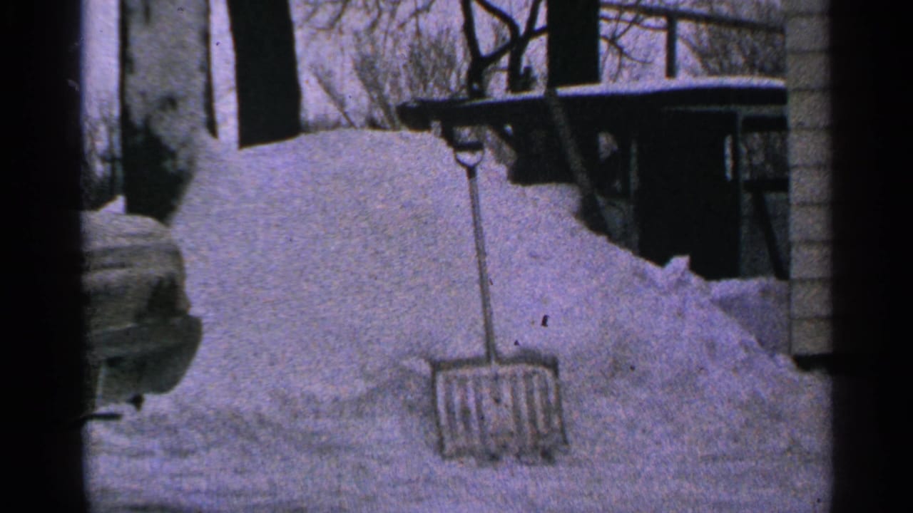 1965 Minnesota 8mm frame showing a tall snowdrift, an upright shovel, house siding, and bare winter branches in a residential yard.