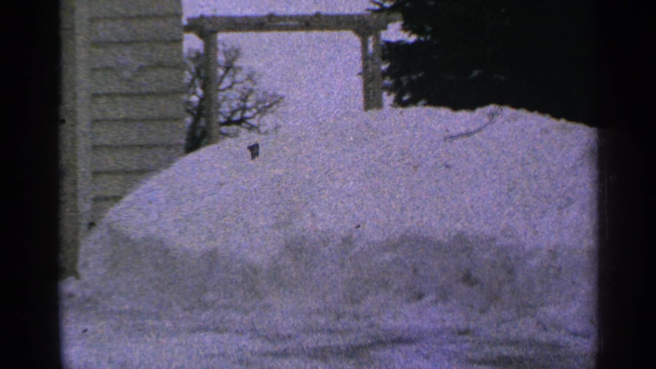 Large snowdrift in a yard with house siding and a wooden frame behind it.