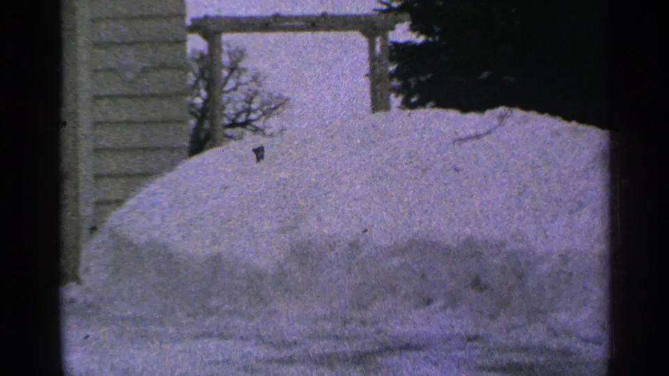 1965 Minnesota 8mm frame with a rounded snowbank, house siding, wooden yard structure, and dark evergreen at the edge.