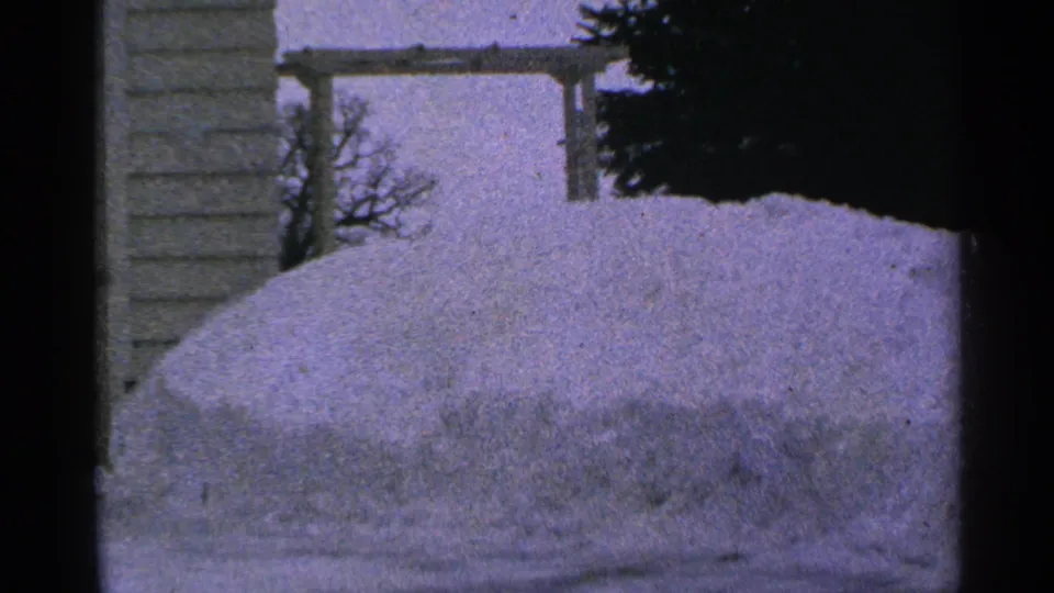 1965 Minnesota 8mm frame showing a high snowbank, house wall, bare trees, and a dark evergreen edge in a quiet winter yard.