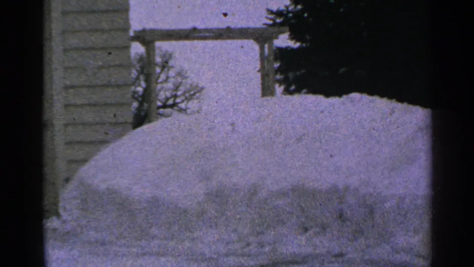 1965 Minnesota 8mm frame of a large yard snowdrift beside house siding, with a simple wooden structure and evergreen in back.