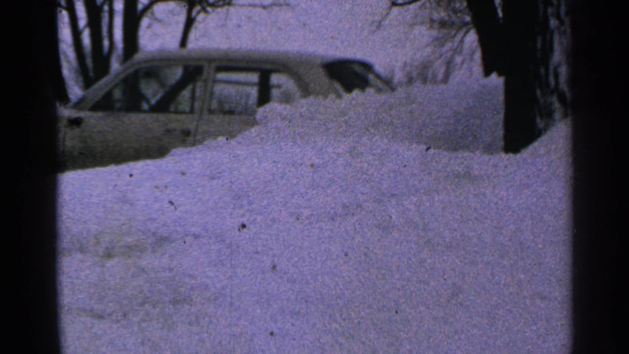Light-colored sedan partly hidden behind a large snowbank near bare trees.