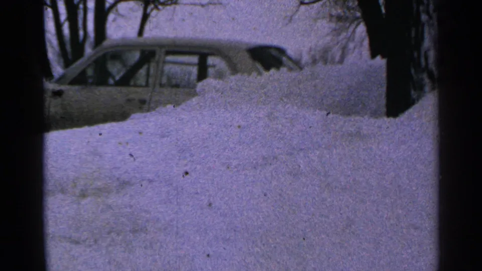 1965 Minnesota winter frame with a parked sedan partly obscured by piled snow in a residential yard.