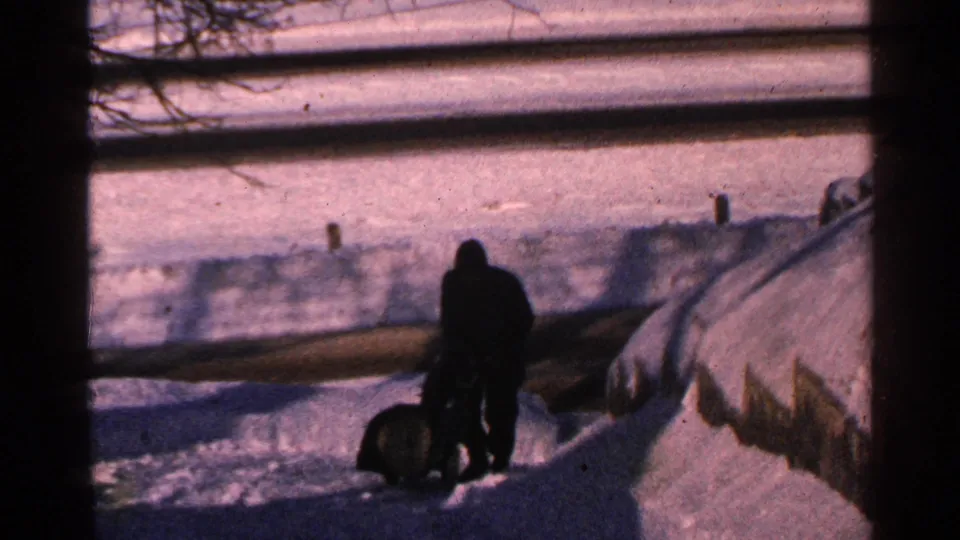 1965 Minnesota home-movie view of a bundled man using a snowblower beside deep snowbanks in a yard.