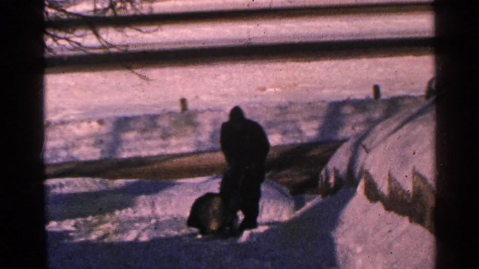 1965 Minnesota winter frame showing a distant bundled man and snowblower near high snowbanks outside a window.