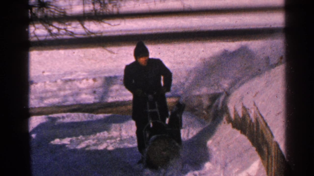 1965 Minnesota 8mm frame showing a bundled adult using a manual snow blower on a snow-covered sidewalk.