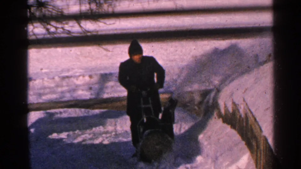 1965 Minnesota 8mm frame showing a bundled adult using a manual snow blower on a snow-covered sidewalk.