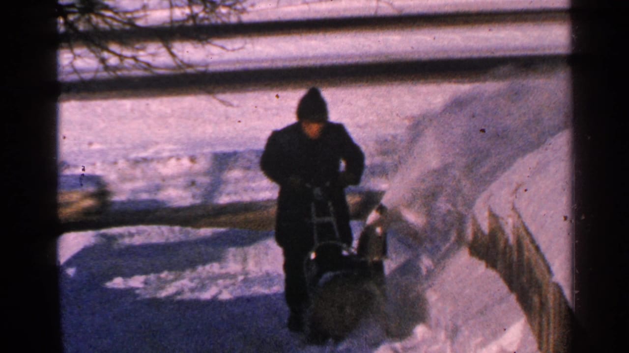 A bundled person operates a manual snow blower on a Minnesota sidewalk, with snow thrown toward a fence.