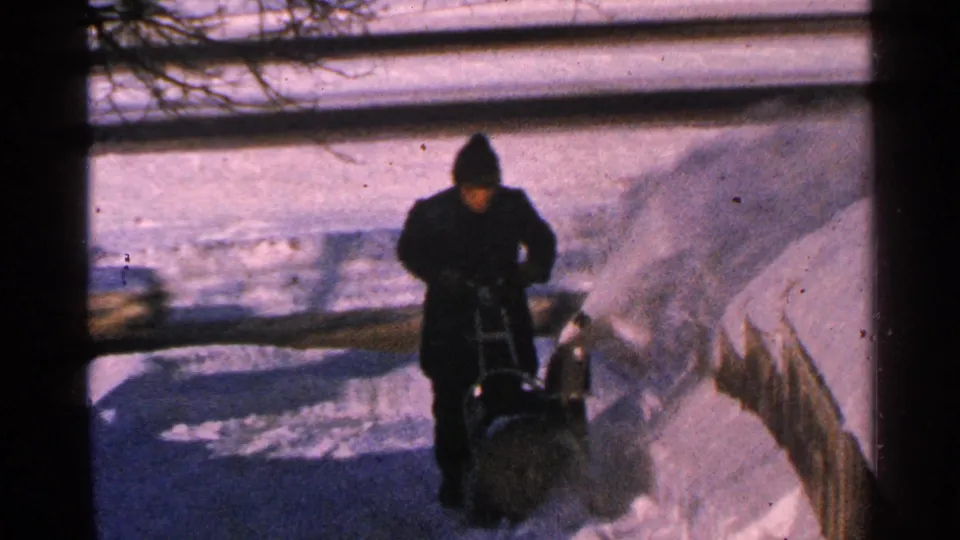A bundled person operates a manual snow blower on a Minnesota sidewalk, with snow thrown toward a fence.