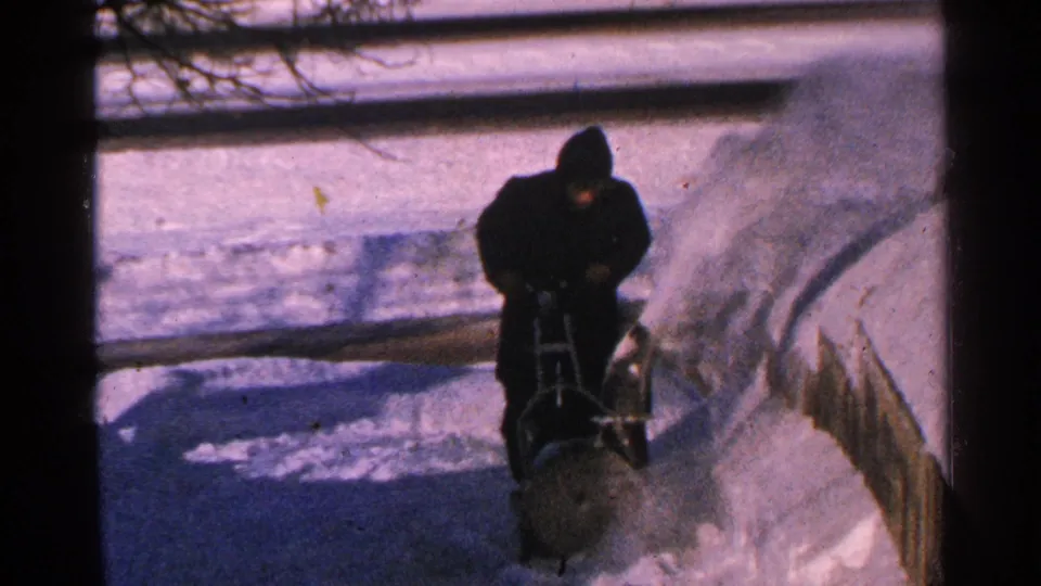 A 1965 home-movie frame shows a bundled adult leaning over a manual snow blower on a winter sidewalk.