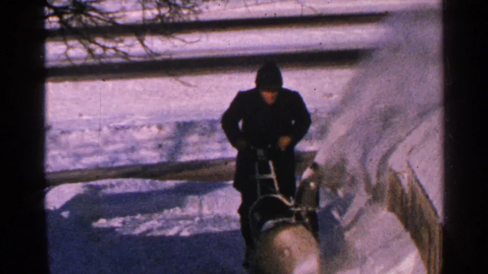 The bundled operator continues clearing a snowy sidewalk in 1965 Minnesota home-movie footage.