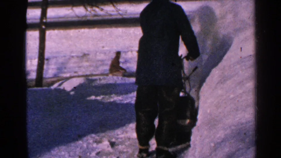 In 1965 Minnesota home-movie footage, a man in a dark snowsuit works near a snowblower beside piled snow and a fence.