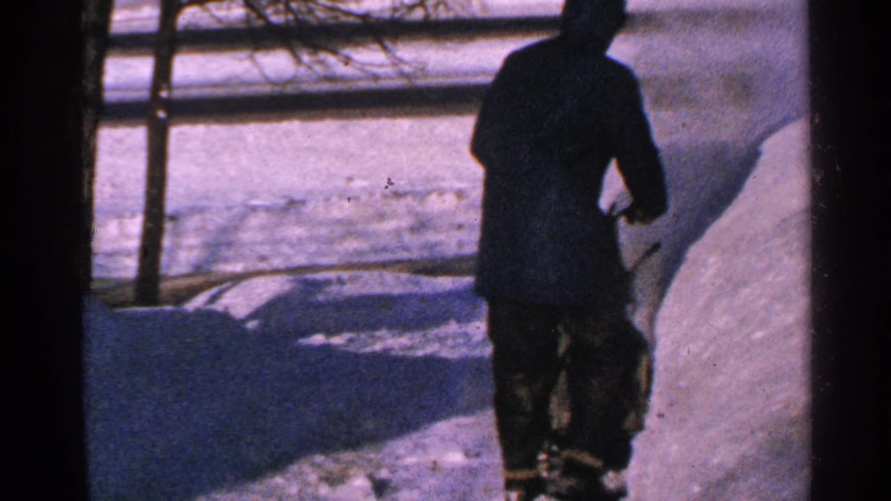 Man in a dark hooded snowsuit walks away through deep snow.