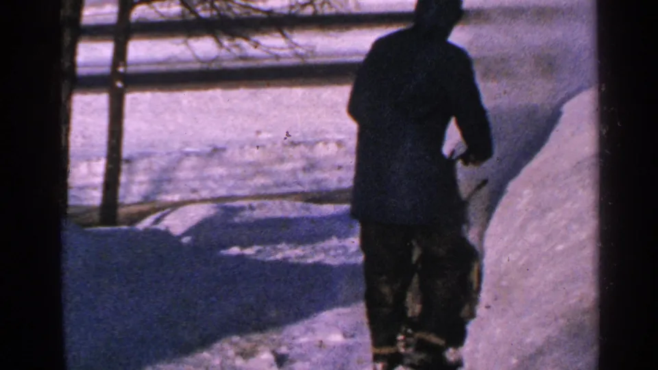 A bundled man in dark winter clothing moves away through deep snow, with a fence and snowbank visible in the background.