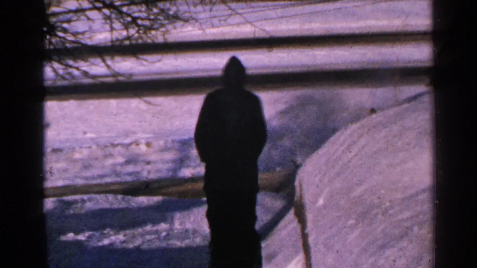 A bundled man faces away beside piled snow in a 1965 Minnesota winter home-movie frame with a fence behind him.