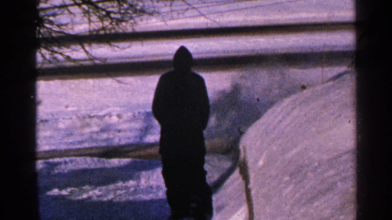 Silhouetted man in a hooded coat stands near a snowy walkway seen through a window frame.