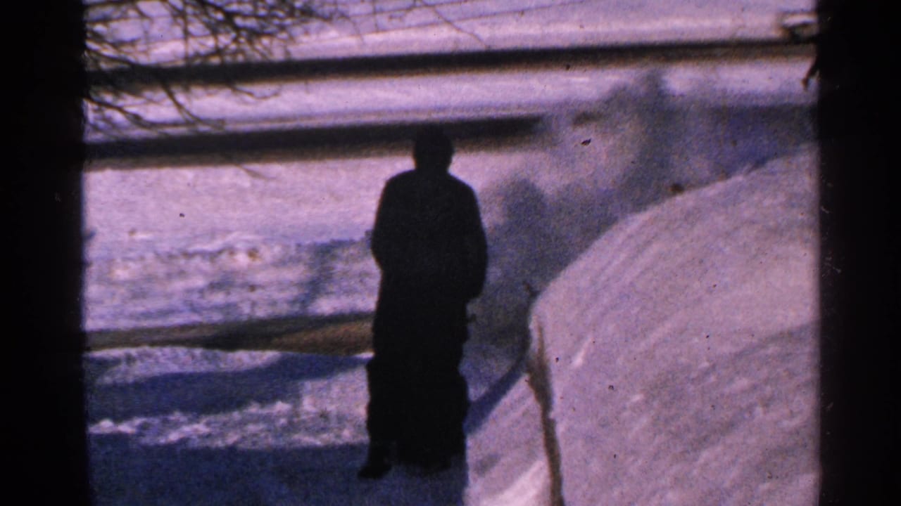 1965 Minnesota home-movie frame with a silhouetted man near a snowbank and plume of blown snow in a winter yard.