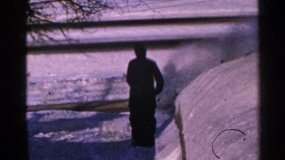 1965 Minnesota home-movie frame with a silhouetted man near a snow-covered walkway and visible blown snow, framed from indoors or behind a window.