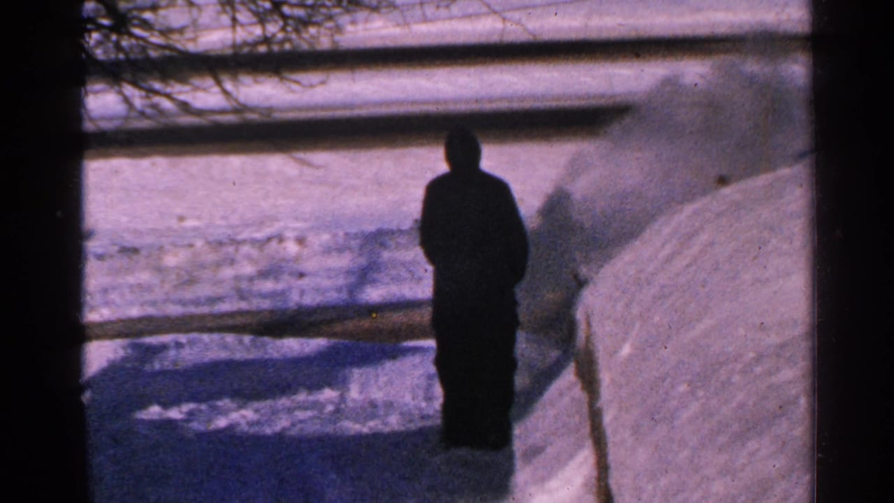 Bundled man stands near deep snow as snow blows across a yard.