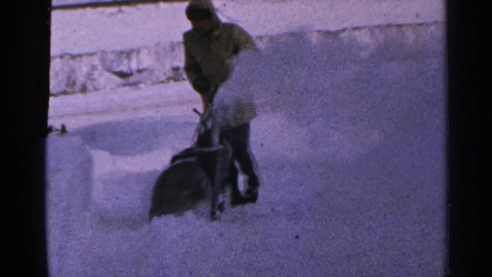 1965 Minnesota home-movie view of a man pushing a snow blower forward through a narrow snowy path.