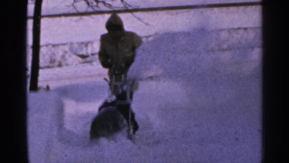 1965 Minnesota frame showing a man behind a snow blower with a large plume of snow arcing across the yard.